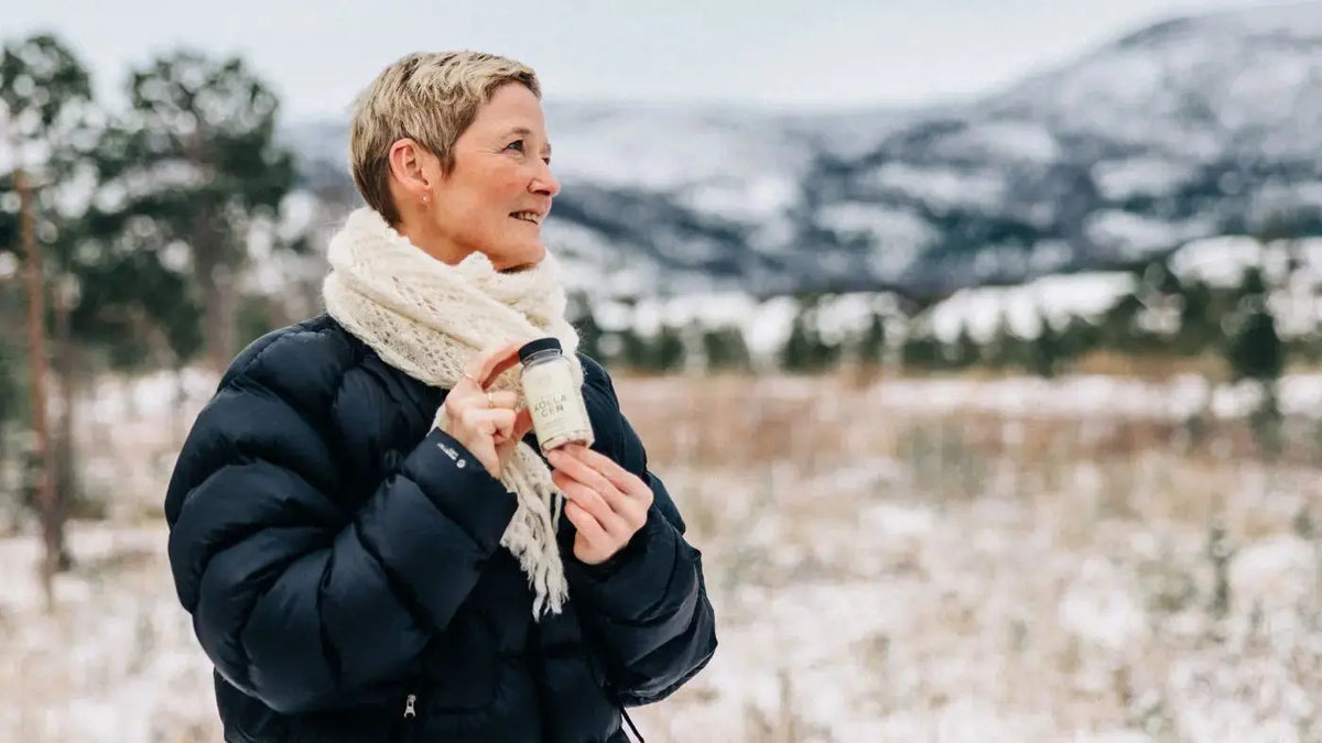 En smilende dame med blondt, kort hår og en svart boblejakke står ute i vinterlandskap med en gul boks Kollagen i hendene.