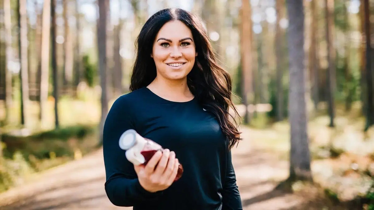 En smilende dame med langt svart hår og en mørkeblå genser står midt i en solfylt skog. Hun vises fra midjen og opp, og i hånden holder hun en boks med kollagen