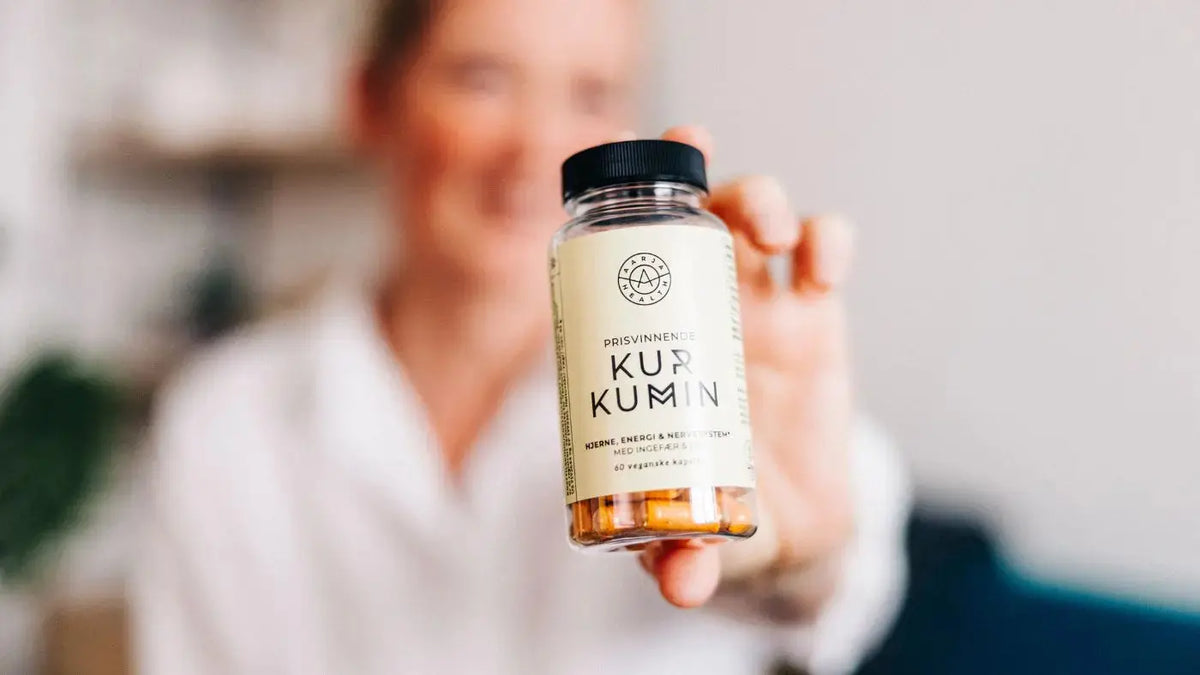 En smilende dame med blondt, kort hår holder en gul boks med kurkumin. Hun er i bakgrunnen og er diffus.